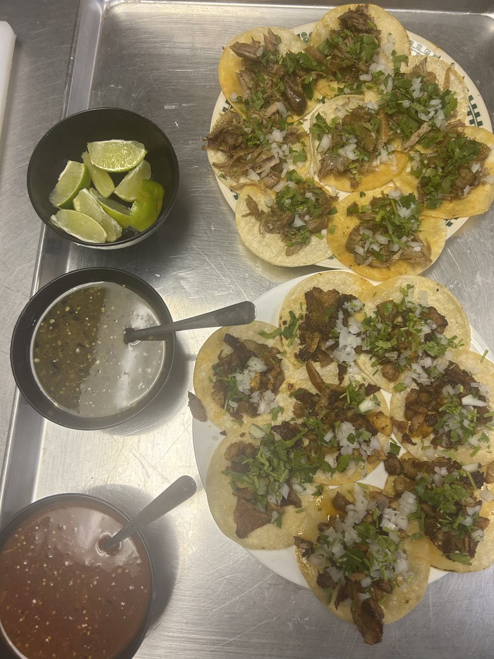 Two plates of tacos with onion and cilantro, salsa verde, salsa roja and limes